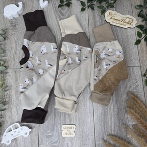 Könnte beinhalten: Drei Babyhosen mit einem Möwenmuster in Braun-, Beige- und Grautönen. Jede Hose hat eine andere Bündchenfarbe, darunter Braun, Beige und Hellbraun. Die Hosen werden auf einer Holzoberfläche mit dekorativen Elementen präsentiert.