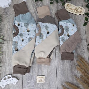 Könnte beinhalten: Drei Babyhosen in Hellblau und Beige. Die Hosen haben ein Heißluftballonmuster und braune Bündchen. Der Text "Traum Nadel" ist auf einem Holzschild zu sehen. Der Text "Schnitt by Lybstes" ist ebenfalls sichtbar.
