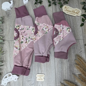 Könnte beinhalten: Drei Babyhosen in verschiedenen Größen mit floralem Muster und mauvefarbenen Akzenten. Die Hosen haben mauvefarbene Bündchen und eine Tasche. Der Hintergrund enthält dekorative Elemente und den Text "Traum Nadel" und "Schnitt by Lydstes".