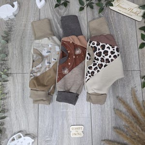 Könnte beinhalten: Drei Babyhosen mit verschiedenen Mustern und Farben. Ein Paar hat einen Möwen-Print, ein anderes ein Bären-Design und das dritte einen Leoparden-Print. Jedes Paar hat ein neutralfarbenes Bündchen und wird auf einer Holzoberfläche präsentiert.