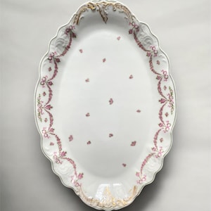 Può includere: Piatto da portata ovale in porcellana bianca con bordo smerlato. Il bordo è decorato con ghirlande e fiocchi floreali rosa, con accenti dorati. Piccoli gruppi di fiori rosa sono sparsi al centro del piatto.