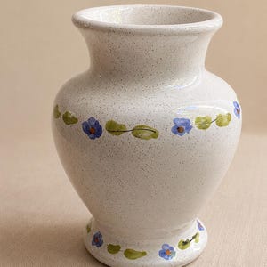 May include: A ceramic vase with a speckled off-white glaze featuring a hand-painted horizontal band of small blue flowers and green leaves around the center and a matching floral pattern around the base, with a flared rim and a rounded body.