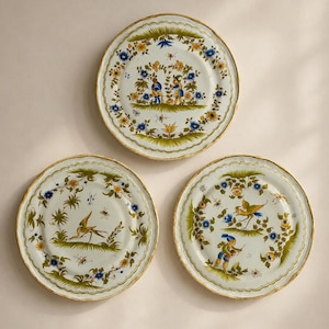 Könnte beinhalten: Drei runde Keramikteller mit gewelltem Rand, verziert mit handgemalten Blumenmotiven in Blau, Gelb und Grün. Die Designs zeigen Figuren in traditioneller Kleidung sowie Vögel inmitten von Blattwerk auf einem weißen glasierten Untergrund.
