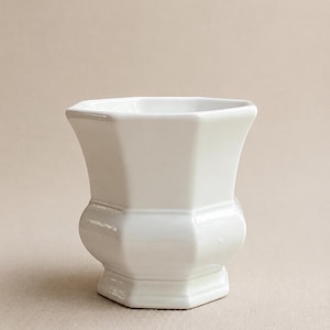 May include: A white ceramic planter with an octagonal shape, featuring a flared rim, a tapered midsection, and a sturdy base. The vessel has a smooth, glossy finish and a clean, geometric design that provides a neutral look for various plants.