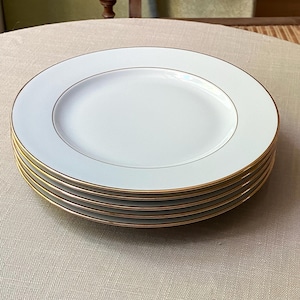 Può includere: Una pila di cinque piatti rotondi in ceramica bianca, ognuno caratterizzato da una finitura liscia e lucida e da un sottile bordo in oro metallizzato lungo il perimetro esterno. I piatti sono uniformi per dimensioni e design.