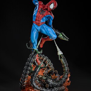 May include: A detailed figurine of Spider-Man in a dynamic pose, clad in a red and blue suit, with a black spider emblem. The figure is mounted on a base featuring mechanical tentacles and a fiery, orange-red glow.