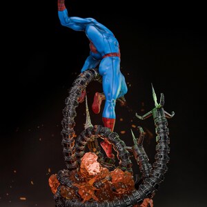 May include: A dynamic statue of Spider-Man in a classic blue and red suit, posed mid-action. He is entangled with mechanical tentacles, and the base features fiery orange and black elements. The statue has a detailed design and is set against a dark background.