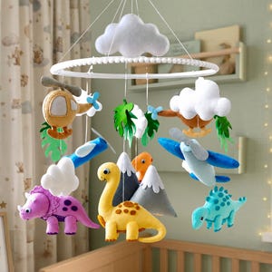 May include: A hanging nursery mobile featuring felt plush figures including a yellow dinosaur, a purple triceratops, a blue dinosaur, a helicopter, an airplane, grey mountains, and white clouds suspended from a circular white frame.