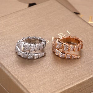 Puede incluir: Dos anillos envolventes con un diseño segmentado similar a una serpiente. Un anillo es de metal plateado con piedras pavé transparentes y el otro es de metal en tono oro rosa con piedras pavé transparentes. Patrón de escamas superpuestas.