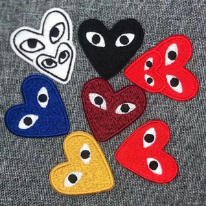 May include: A selection of heart-shaped embroidered patches in diverse colours, such as white, black, red, blue, and yellow. Each patch displays a distinctive eye design. The patches are arranged on a grey fabric surface.