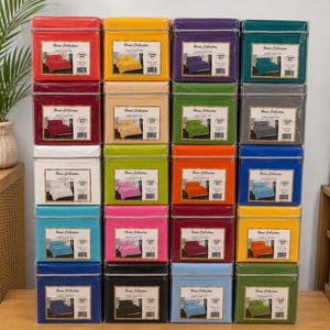 May include: A stack of twenty colorful sheet set boxes. The boxes are in various colors, including red, yellow, purple, teal, green, tan, gray, white, orange, pink, blue, and black. Each box has a label that reads "Home Collection 4 Piece Sheet Set".