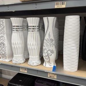 Könnte beinhalten: Eine Sammlung dekorativer Vasen in verschiedenen Stilen und Farben. Die Vasen sind weiß, schwarz und silber. Einige haben strukturierte Oberflächen, während andere florale oder geometrische Designs aufweisen. Die Vasen werden auf einem Regal ausgestellt.