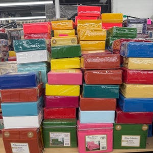 May include: A display of colorful bed sheet sets in various colors, including red, yellow, green, and blue. The sets are neatly stacked and wrapped in clear plastic, with some boxes visible at the bottom.