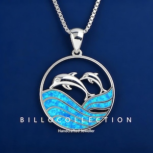 May include: Silver pendant necklace with a circular design depicting two dolphins above blue waves. The pendant has a blue opal inlay and hangs from a silver chain. The text "BILLO COLLECTION" and "Handcrafted Jeweller" are visible.