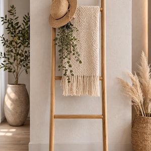 May include: A wooden ladder with a straw hat, a cream-colored fringed blanket, and hanging greenery. The ladder is leaning against a neutral-colored wall. A potted plant and a basket with tall grass are also visible.