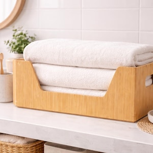 May include: A wooden organizer holds folded white towels on a bathroom countertop. The organizer is light brown and has a cutout handle on each side. A round mirror and a small plant are in the background.