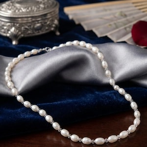 Peut inclure: Un collier de perles délicat avec des perles ovales et de petites perles transparentes. Le collier est présenté sur un tissu de satin argenté sur une surface de velours bleu foncé. Une boîte à bijoux argentée et un éventail décoratif sont en arrière-plan.