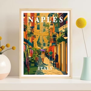 May include: A framed art print featuring a vibrant street scene in Naples, Italy, with warm yellow buildings, a large gold bow decoration, and people walking on a cobblestone path. The text NAPLES and ITALY is printed in white capital letters.