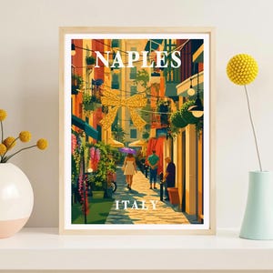 May include: A framed travel poster featuring a vibrant street scene in Naples, Italy, with the text NAPLES ITALY in white. The illustration depicts a narrow cobblestone alleyway with colorful buildings, hanging plants, a large gold bow, and pedestrians.