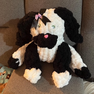 May include: A handmade plush dog with a black and white color scheme. The dog has black floppy ears, a black bow with a pink center, and green button eyes. The dog's nose is a small pink heart.