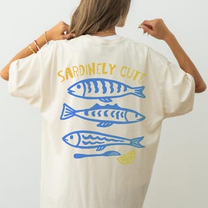May include: Cream-colored t-shirt with the text "Sardinely Cute" in yellow and blue. The shirt features three blue fish illustrations with a lemon slice and a fork. The shirt is a casual, oversized style.