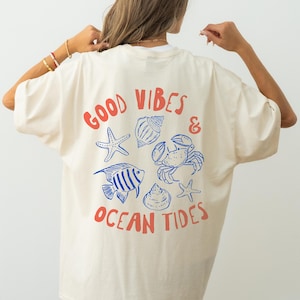 May include: Cream-coloured t-shirt with the words "GOOD VIBES & OCEAN TIDES" in red and blue. The shirt features illustrations of a starfish, seashells, a crab, and a fish. The shirt is a relaxed fit.