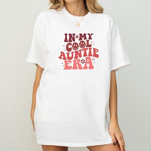 Könnte beinhalten: Weißes T-Shirt mit dem Text "IN MY COOL AUNTIE ERA" in einem Retro-Schriftstil. Das Design beinhaltet ein lächelndes Gesicht, Blumen und Sterne in Rot- und Rosatönen. Das Shirt hat einen lässigen, übergroßen Stil.