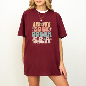 Könnte beinhalten: Burgunderfarbenes T-Shirt mit dem Text "IN MY SOUR DOUGH ERA" in einem Retro-Schriftstil. Das Design beinhaltet Schmetterlinge und Blumen in Pastellfarben. Das T-Shirt hat kurze Ärmel und eine lockere Passform.