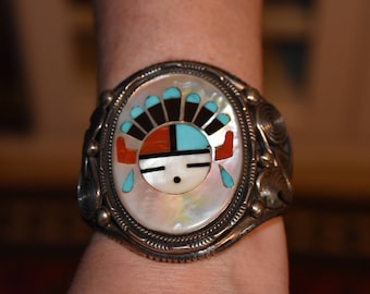 May include: A silver bracelet featuring a round, white and turquoise inlay depicting a Native American face. The face is surrounded by a black and red design.