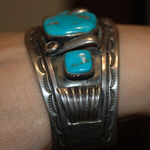 May include: A silver bracelet with turquoise stones set in a geometric design. The bracelet is worn on a person's wrist.