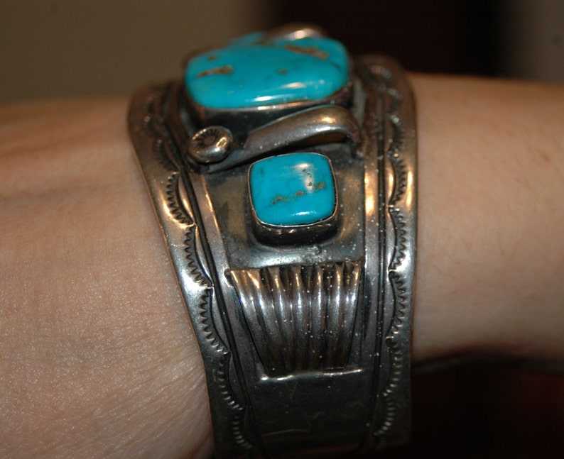 May include: A silver bracelet with turquoise stones set in a geometric design. The bracelet is worn on a person's wrist.