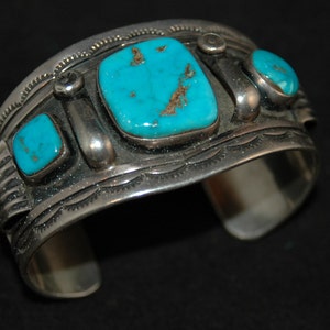 May include: A silver cuff bracelet featuring turquoise stones. The bracelet has an intricate design with a repeating pattern.