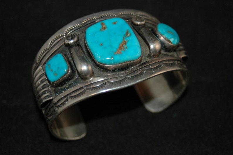 May include: A silver cuff bracelet featuring turquoise stones. The bracelet has an intricate design with a repeating pattern.