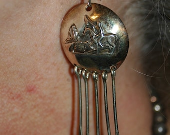 May include: Silver dangle earrings with a round charm featuring a detailed embossed design of a horse and rider. The charm is attached to a series of dangling silver chains.