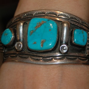 May include: A silver bracelet with turquoise stones. The bracelet features a decorative design and is worn on a person's wrist.