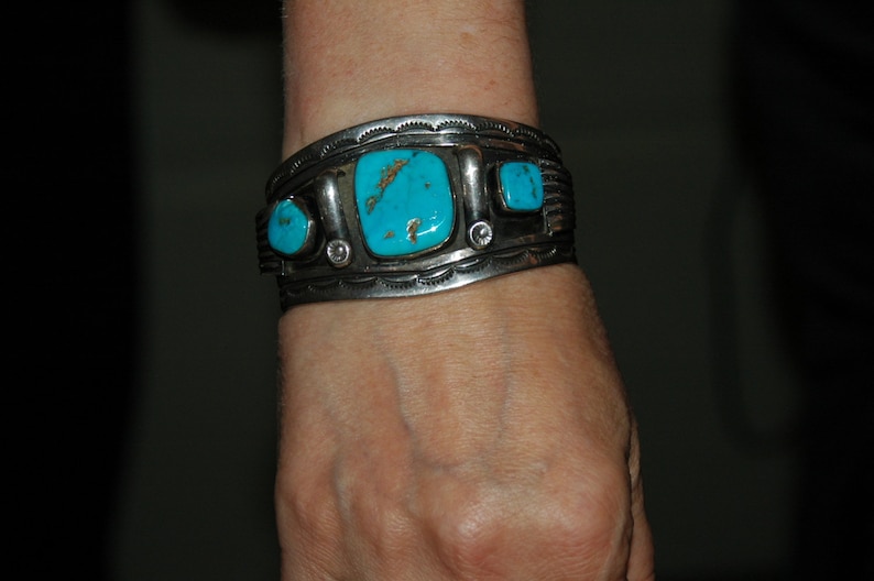 May include: A silver bracelet with turquoise stones set in a silver band. The bracelet is worn on a person's wrist.