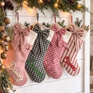 May include: Four personalised Christmas stockings hanging in a row. The first is solid dusty pink with the name Daisy, the second is black and white plaid with the name Lauren, the third is red and white gingham with the name Sophia, and the fourth is red and white striped with the name Owen. Each stocking features a fabric bow at the top.