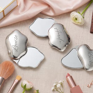 May include: Three silver-toned, irregular-shaped compact mirrors with smooth, reflective surfaces. Each mirror is engraved with a name in cursive script: Louise, Amyle, and Becky. The mirrors are hinged, allowing them to open to reveal a reflective glass interior.