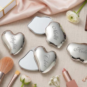 May include: Four silver-toned compact mirrors with irregular, organic shapes and heart-like silhouettes. Each mirror is engraved with a name in elegant script: Sophia, Molly, Clar, and Helen. The mirrors have a polished, reflective metallic finish.