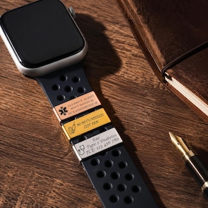 May include: A black smartwatch with a perforated silicone strap featuring three metallic medical alert charms. The charms are rose gold, gold, and silver, engraved with medical information including allergy alerts, medication warnings, and contact details.