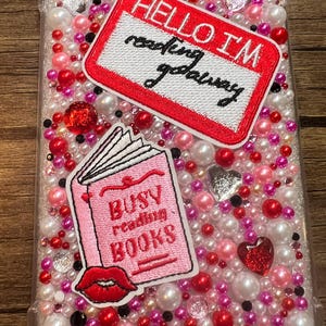 May include: Clear phone case adorned with white, pink, and red beads. The case has two embroidered patches: one reads "HELLO I'M reading getaway" and the other says "BUSY reading BOOKS" with a red lip graphic.