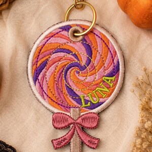 May include: A circular embroidered keyring shaped like a lollipop with a swirling pattern of orange, purple, and pink thread. The name LUNA is stitched in lime green letters. A pink embroidered bow sits at the base, attached to a gold metal clasp.