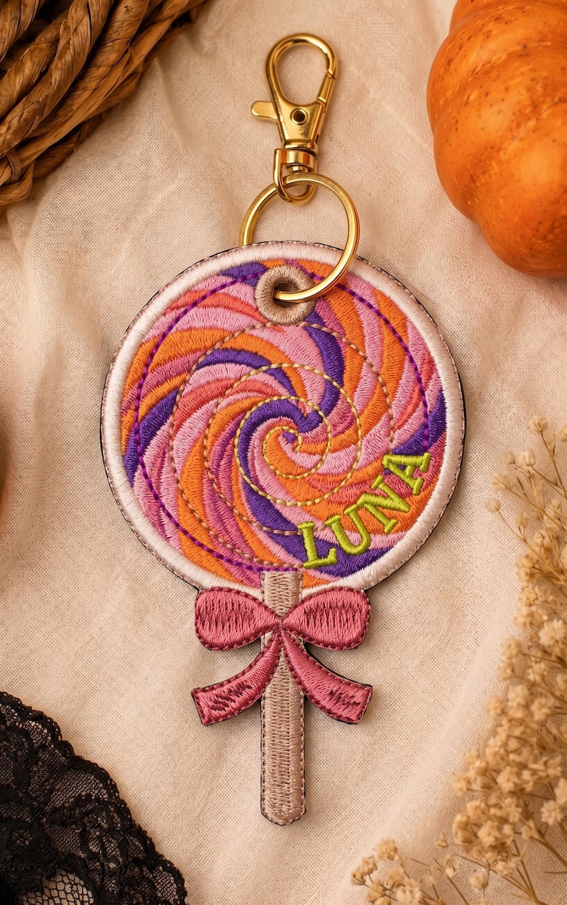 May include: A circular embroidered keyring shaped like a lollipop with a swirling pattern of orange, purple, and pink thread. The name LUNA is stitched in lime green letters. A pink embroidered bow sits at the base, attached to a gold metal clasp.