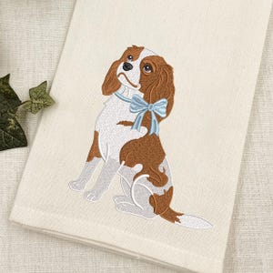 Könnte beinhalten: Ein cremefarbenes Küchentuch mit einer Stickerei eines Cavalier King Charles Spaniels. Der Hund ist weiß und braun, hat lange Ohren und ein hellblaues Band um den Hals. Die Stickerei zeichnet sich durch detaillierte Stiche aus.