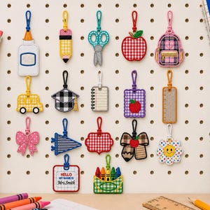May include: A collection of seventeen colorful embroidered keychains featuring school-themed designs including a glue bottle, pencil, scissors, apple, backpack, school bus, graduation cap, notebook, ruler, bow, pennant, and a name tag reading Hello My Name Is Mrs. Smith.