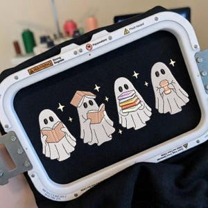 May include: An embroidery hoop holding black fabric with a machine-stitched design of four white ghosts. The ghosts are depicted reading books, holding a stack of colorful books, and holding a coffee cup, accented by small yellow star shapes.