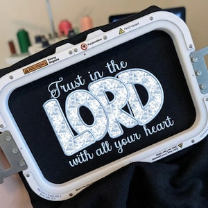 以下が含まれることがあります： 白い長方形の刺繍枠に黒い布が固定されており、Trust in the Lord with all your heartという文字が刺繍されています。LORDという単語は青と白の花柄で、他の単語は白い筆記体で刺繍されています。