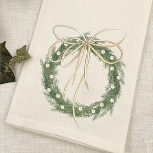 May include: A cream-colored fabric towel featuring a printed illustration of a green pine wreath adorned with small white pearls and a large, flowing beige ribbon tied in a bow at the top, centered on the textile surface.