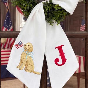 May include: White fabric banner with a green wreath and a large white bow. One side features a golden doodle dog holding an American flag. The other side has a red embroidered letter "J".