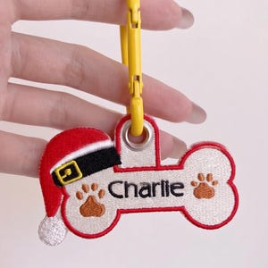 May include: A bone-shaped embroidered keychain featuring a red and white Santa hat, two brown paw prints, and the name Charlie stitched in black thread. The white fabric tag has a red border and is attached to a bright yellow plastic carabiner clip.